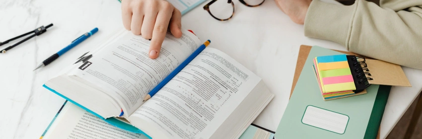 jovem-de-moletom-bege-sentado-a-mesa-cheia-de-cadernos-e-canetas-com-livro-de-estudos-aberto-apontando-para-pagina-lendo-e-segurando-oculos-marrom-com-a-outra-mao-